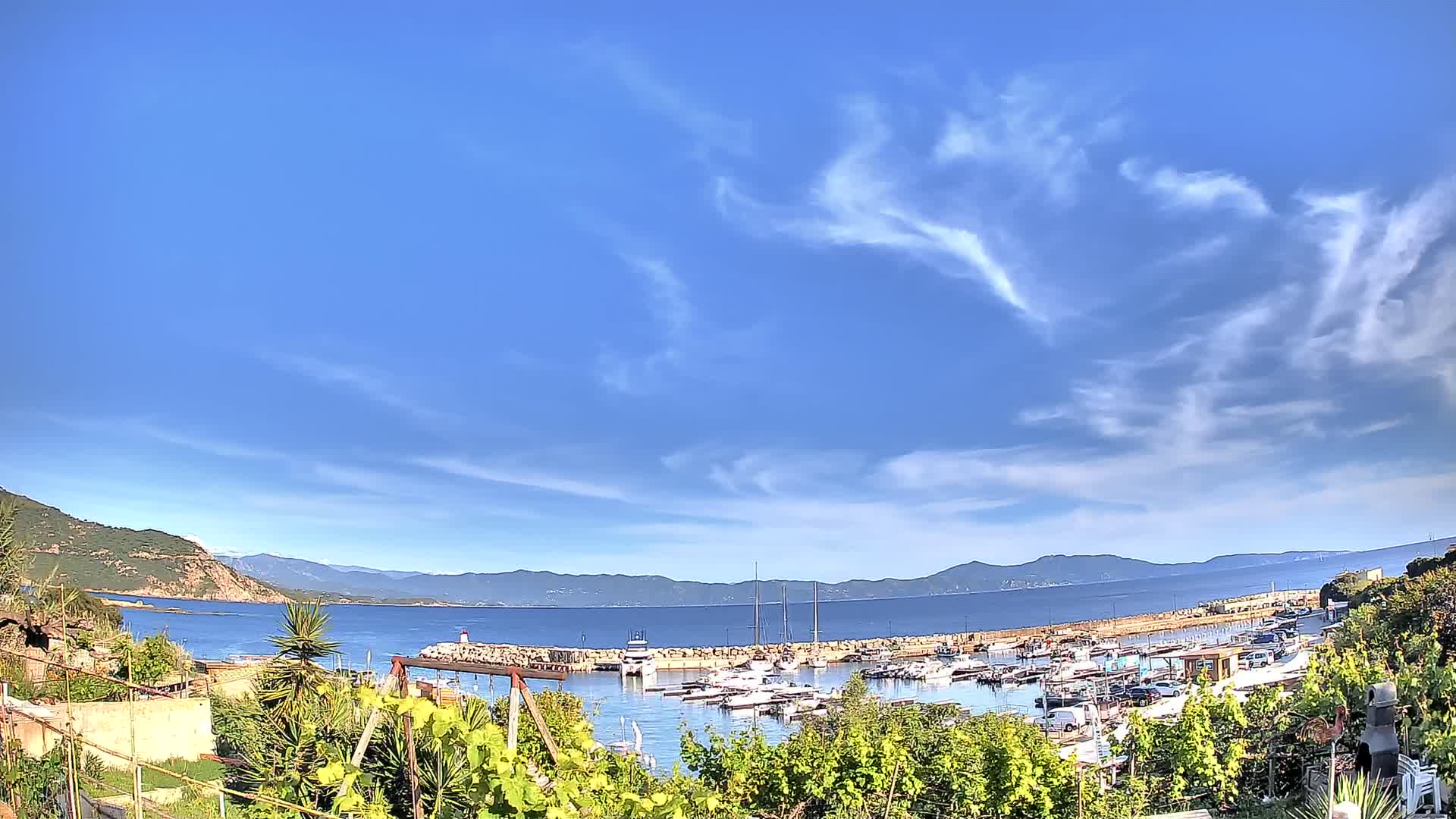 A calm, sunny day reveals a small harbor filled with boats, nestled between hills and a clear blue sea under a partly cloudy sky.