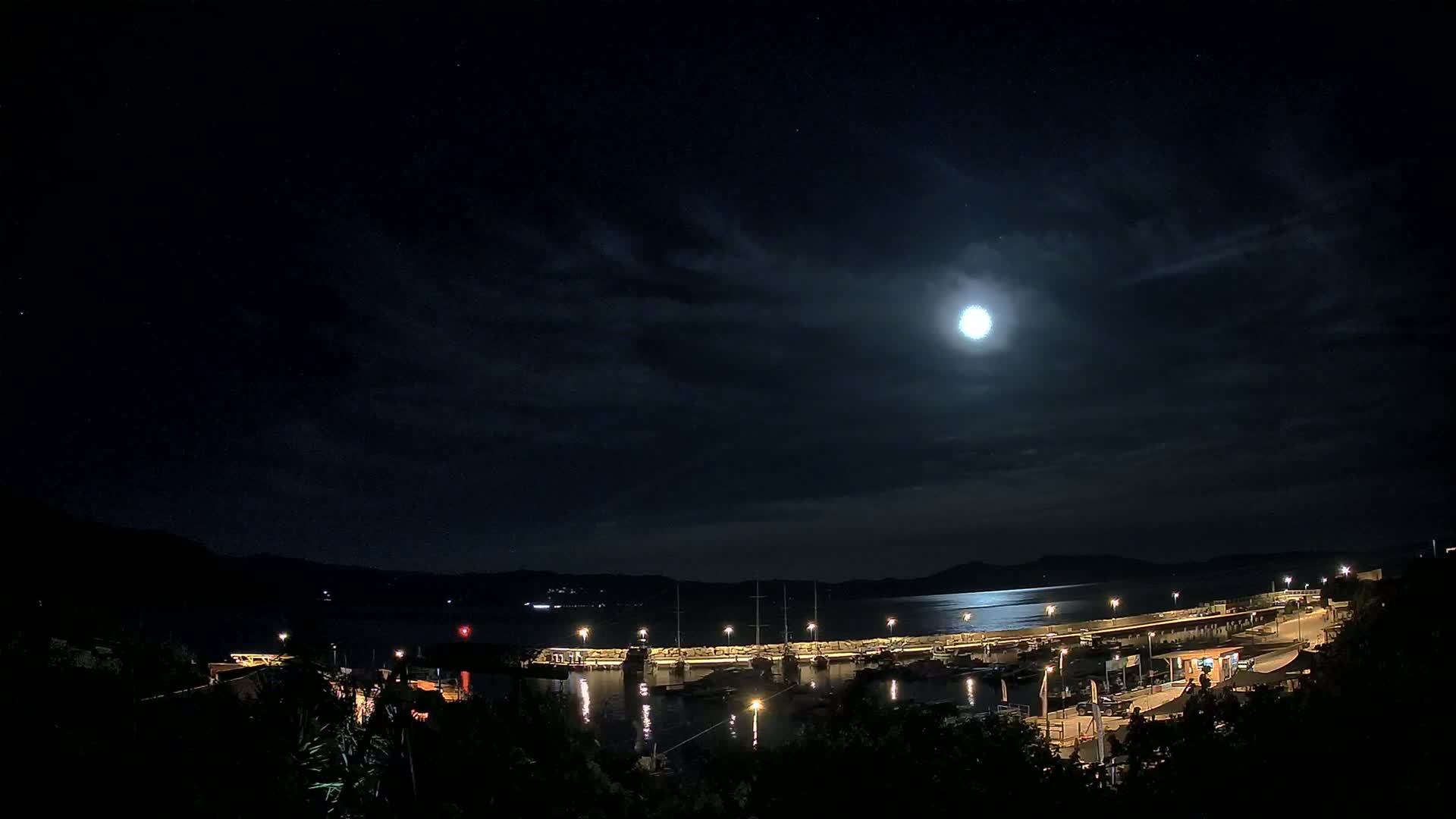 A moonlit night scene shows a harbor filled with boats, illuminated by lights, under a partly cloudy sky.