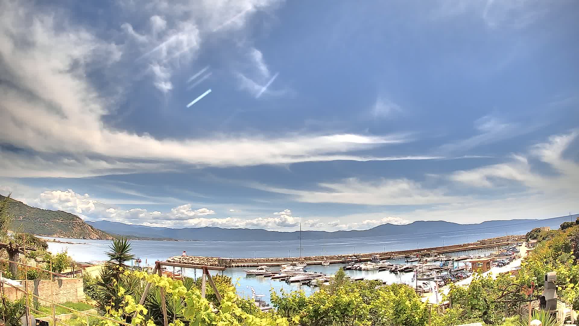 A partly cloudy sky overlooks a calm bay filled with boats, a stone harbor wall, and distant mountains.