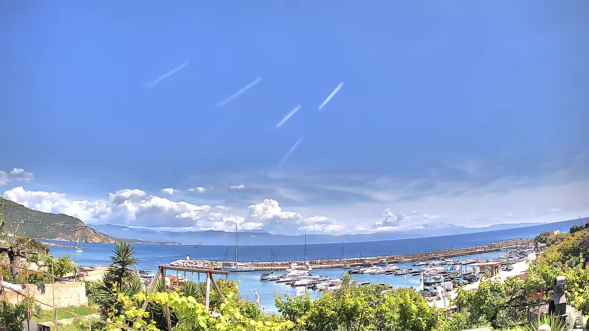 A marina filled with boats is nestled between a rocky coastline and a calm sea under a partly cloudy, sunny sky.