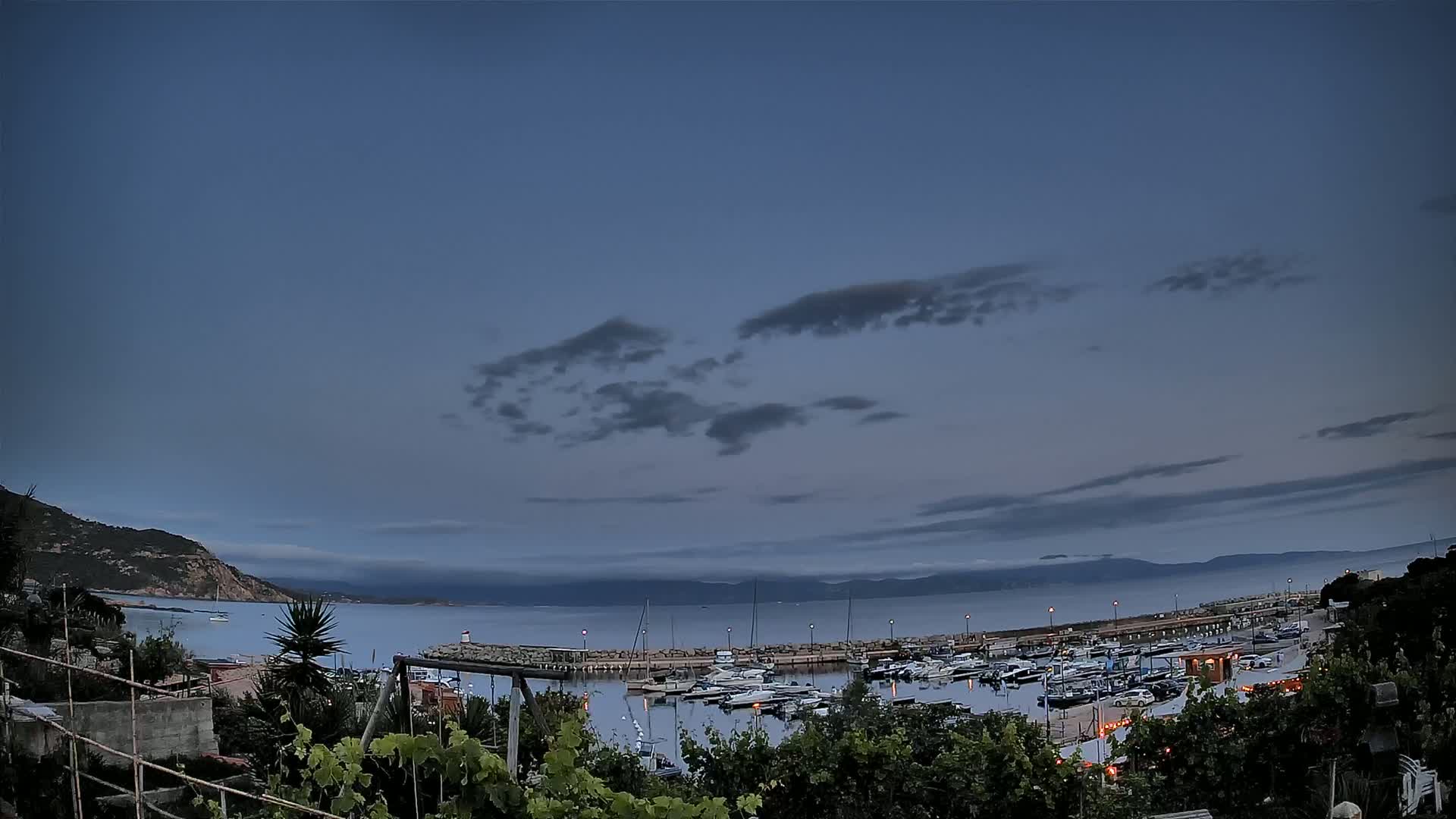 A calm bay filled with moored boats is seen at dusk under a partly cloudy sky from an elevated vantage point, with hills visible in the distance.