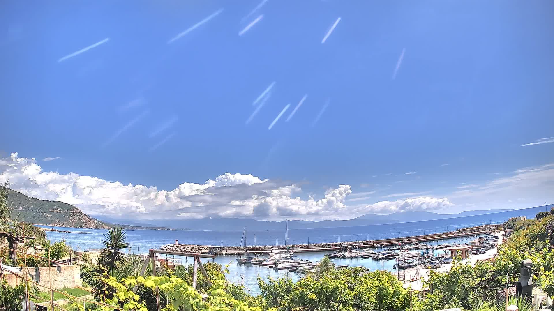 A partly cloudy, sunny day reveals a harbor filled with boats, nestled between a verdant hillside and a calm sea with distant mountains under a mostly blue sky.