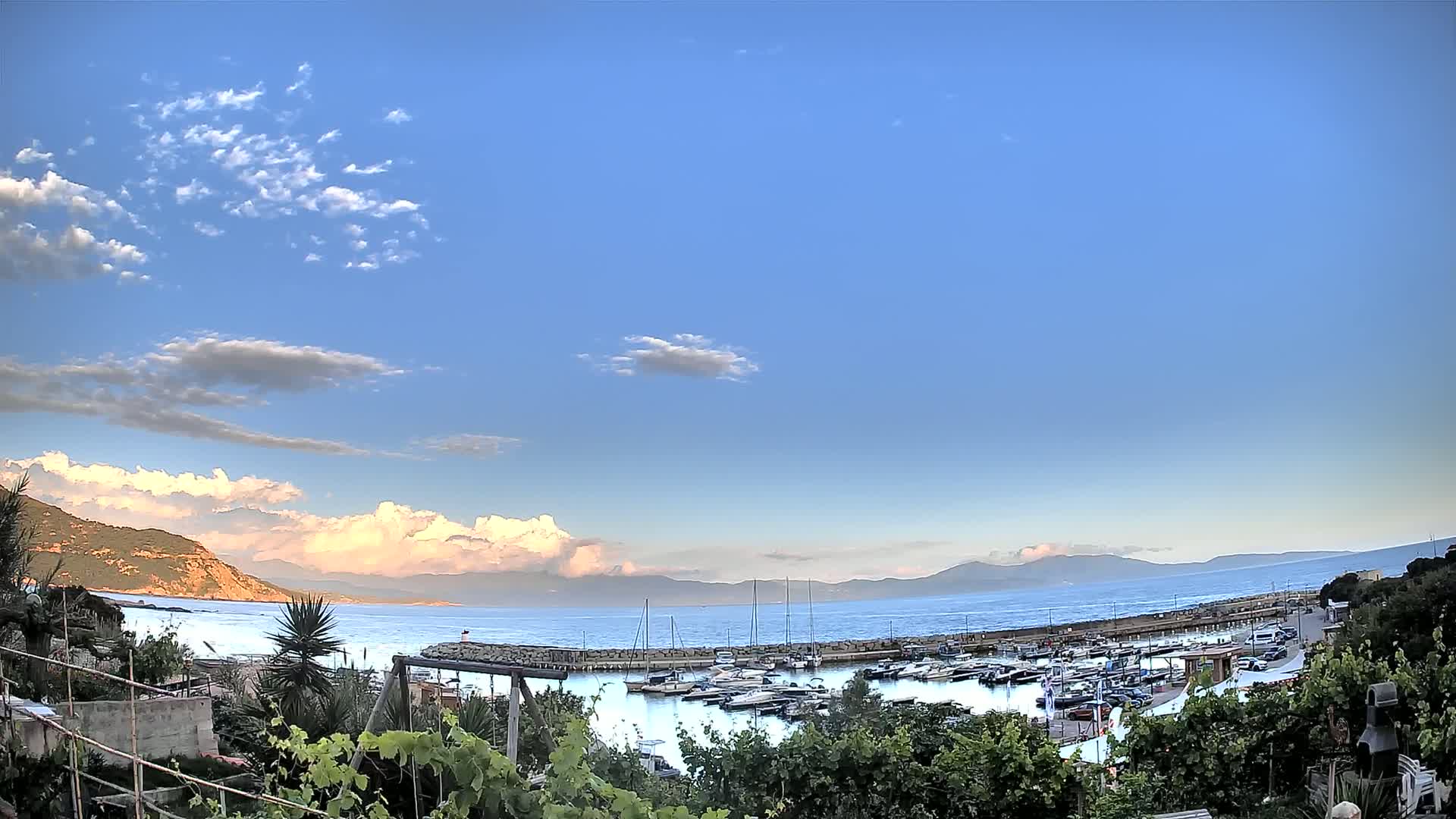 A mostly clear blue sky with scattered clouds overlooks a calm harbor filled with sailboats and motorboats, nestled between a verdant hillside and a distant mountain range.