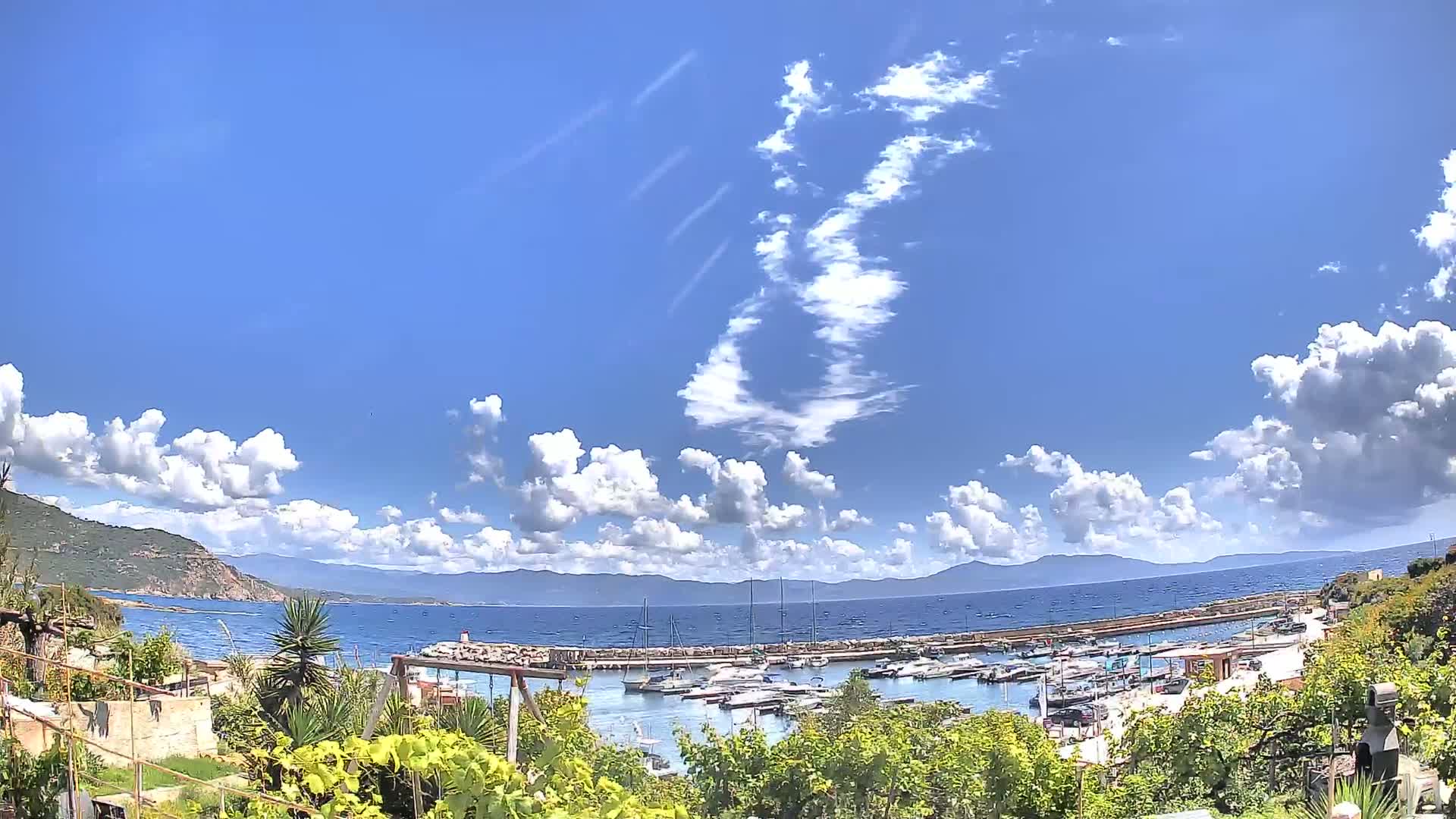 A partly cloudy sunny day overlooks a small harbor filled with boats, situated between green hills and a calm blue sea.