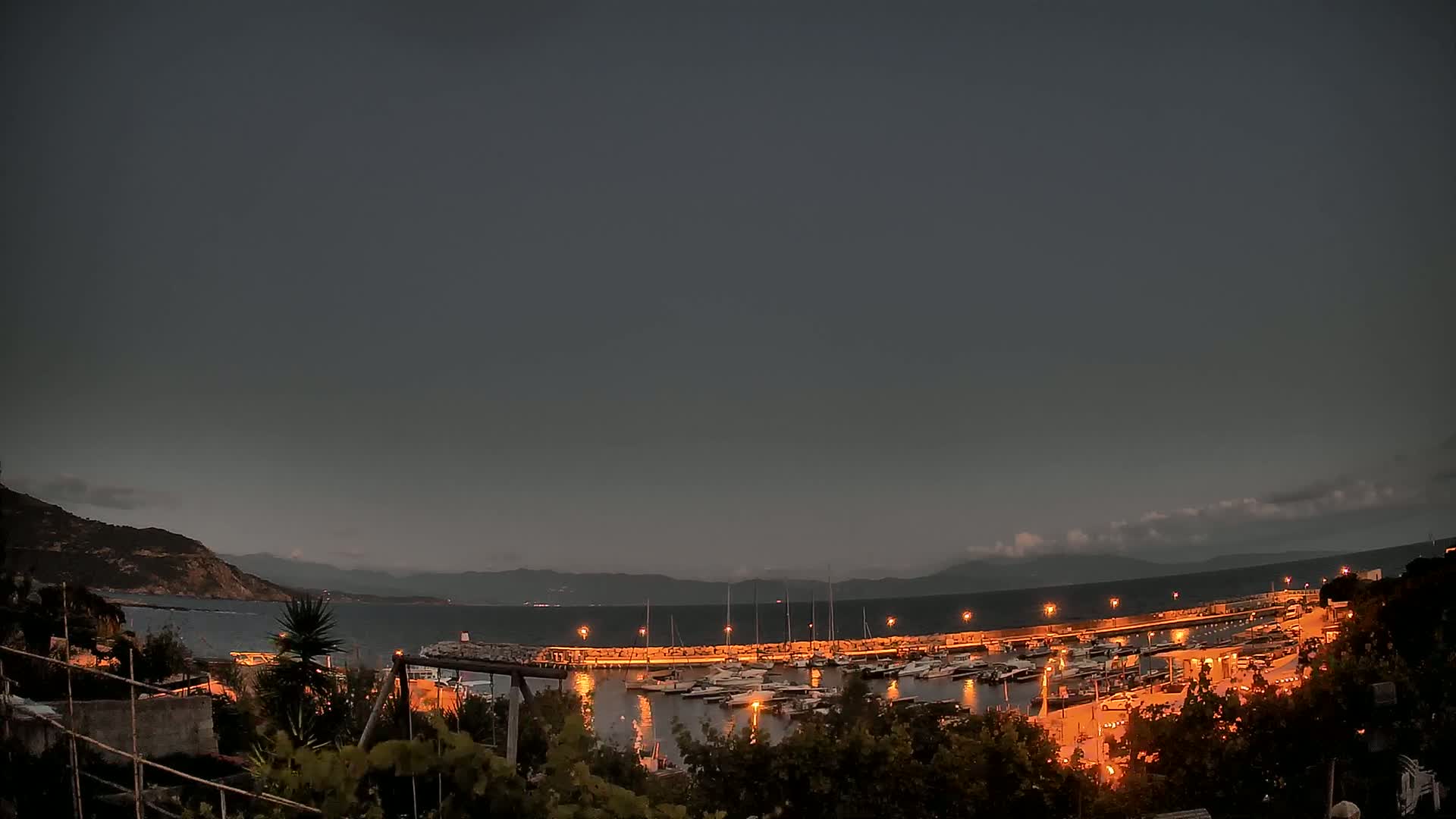 A nighttime view of a harbor filled with boats, nestled between a dark, rocky coastline and a calm sea under a mostly cloudy, dusky sky.