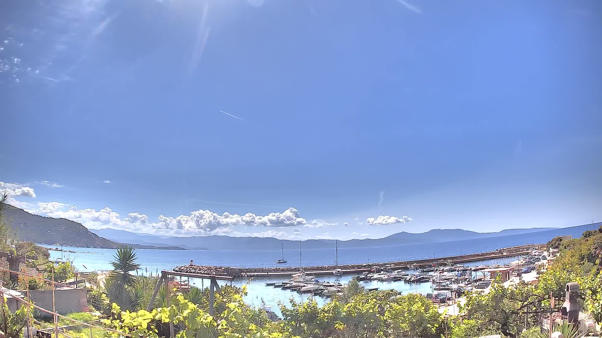 A sunny day reveals a calm bay filled with boats, nestled between a verdant shoreline and distant mountains under a mostly clear sky.