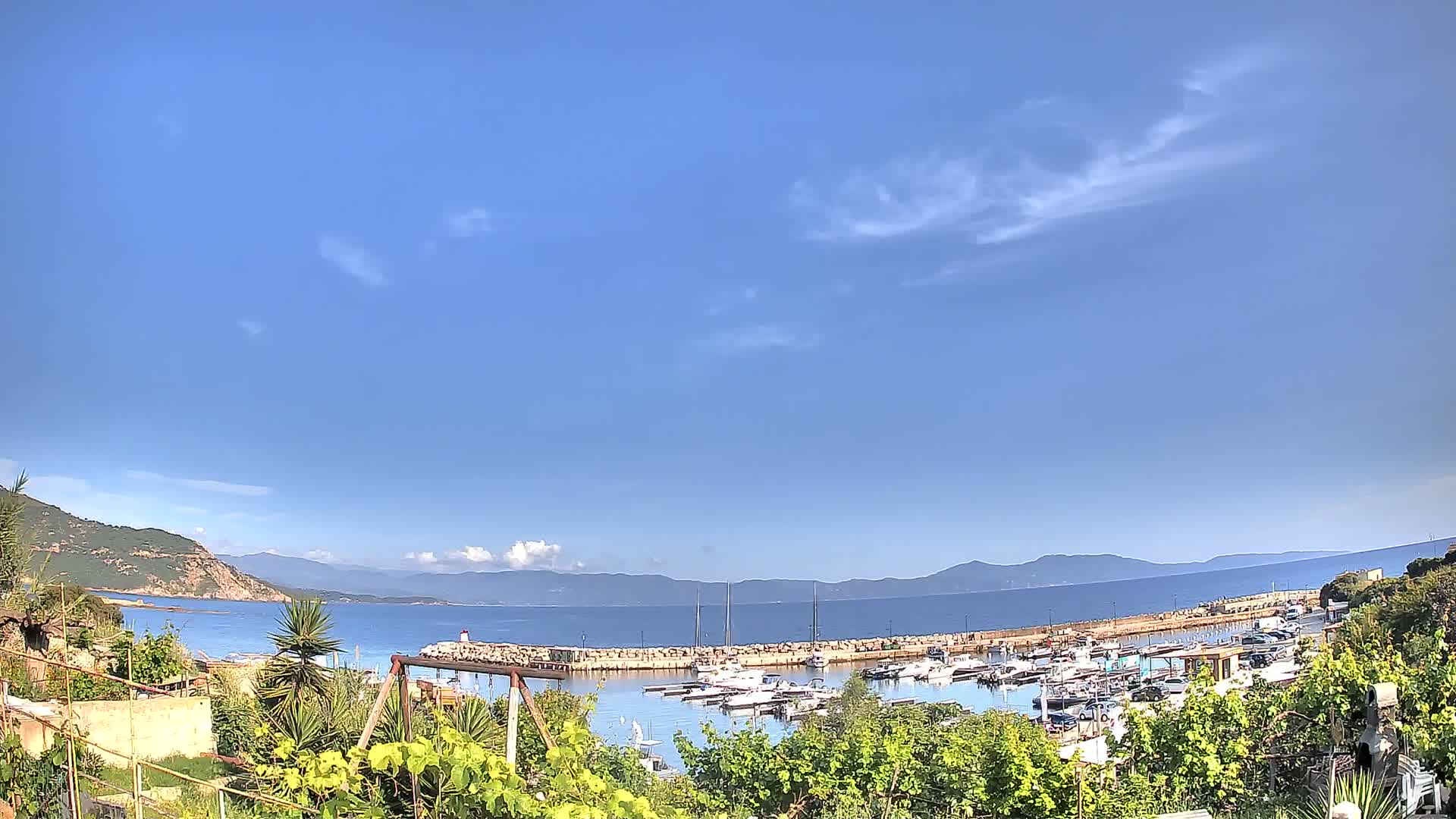 A mostly sunny day shows a calm harbor filled with boats, nestled between a green hillside and distant mountains.