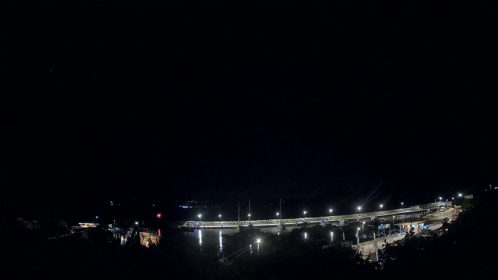A nighttime view of a harbor with boats docked along a lit seawall under a clear, dark sky.