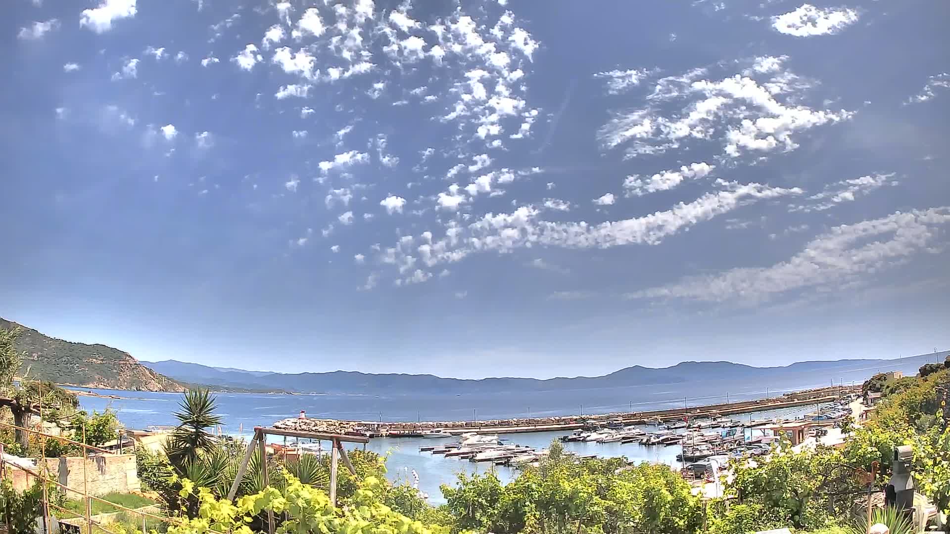 A partly cloudy sky overlooks a calm bay filled with boats, nestled between hills and mountains.