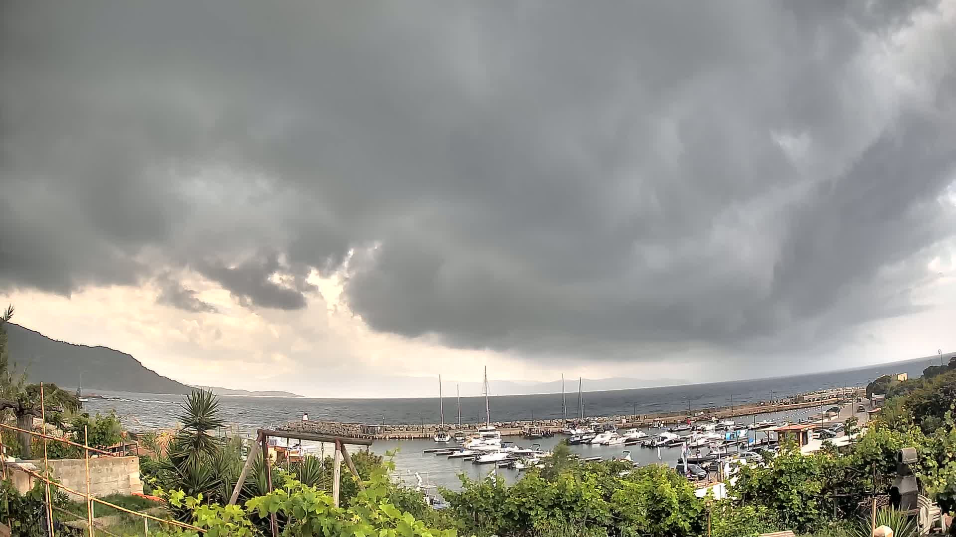 A marina filled with boats is situated on a calm sea under a dark, stormy sky.