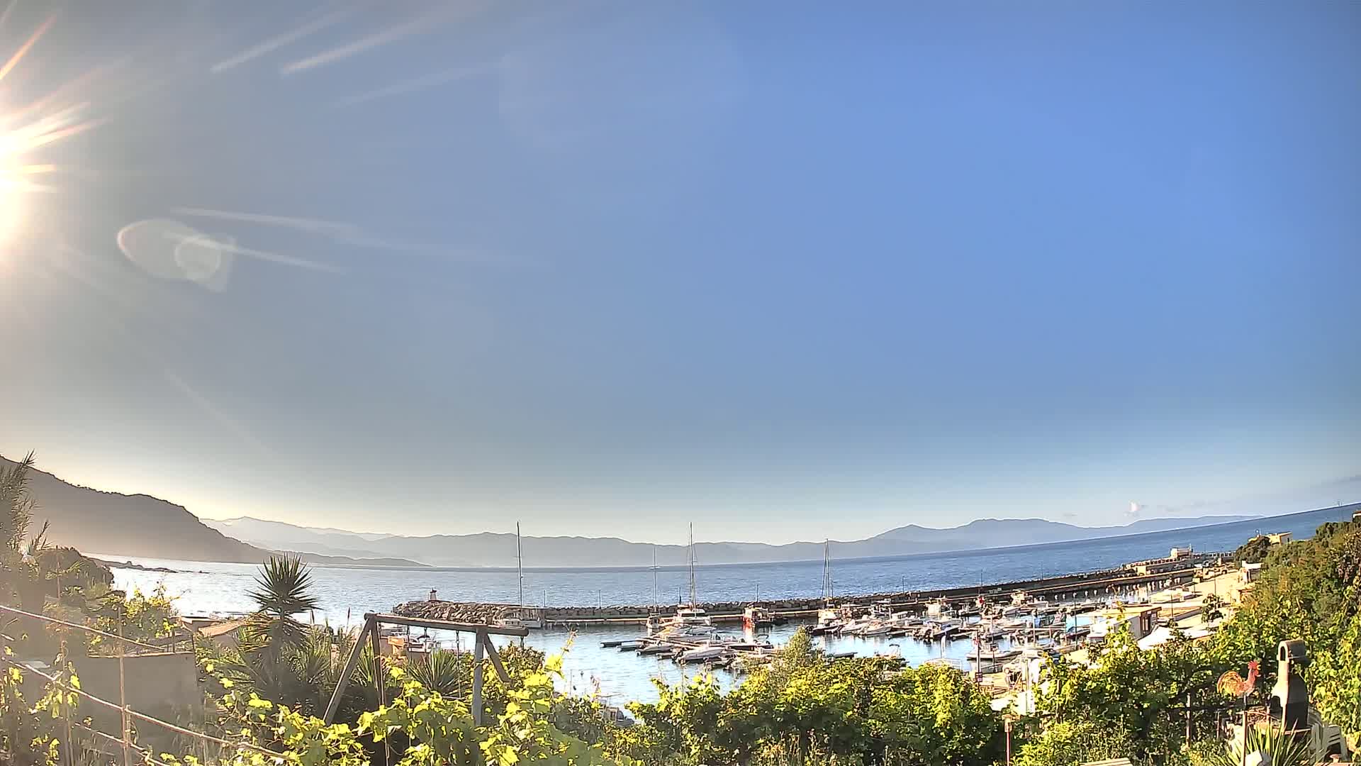 A sunny day reveals a harbor filled with boats, nestled between a calm sea and distant mountains.