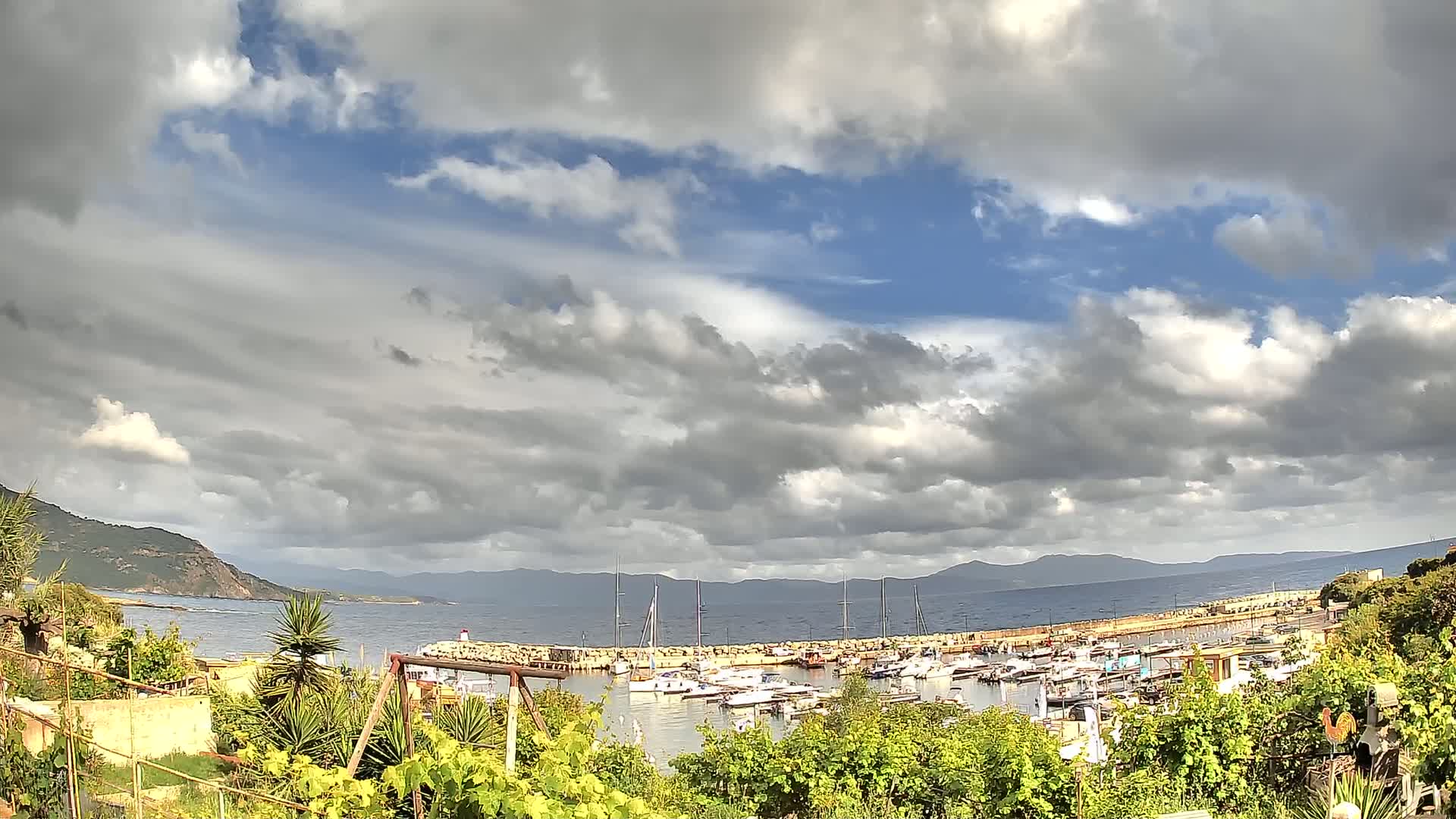 A partly cloudy sky overlooks a calm harbor filled with numerous boats, nestled between a verdant coastline and distant mountains.