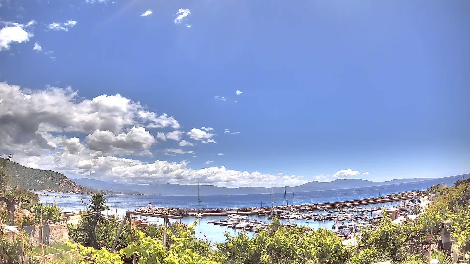 A partly cloudy blue sky overlooks a calm bay filled with moored boats, backed by a mountainous coastline.