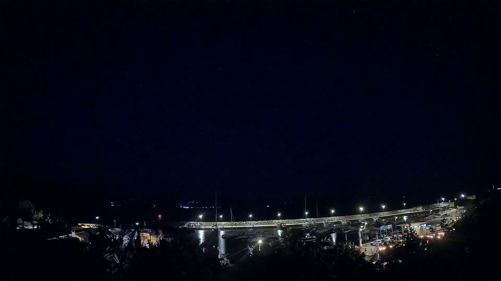 Under a clear, starlit night sky, a marina filled with boats is illuminated by lights along a curved pier.