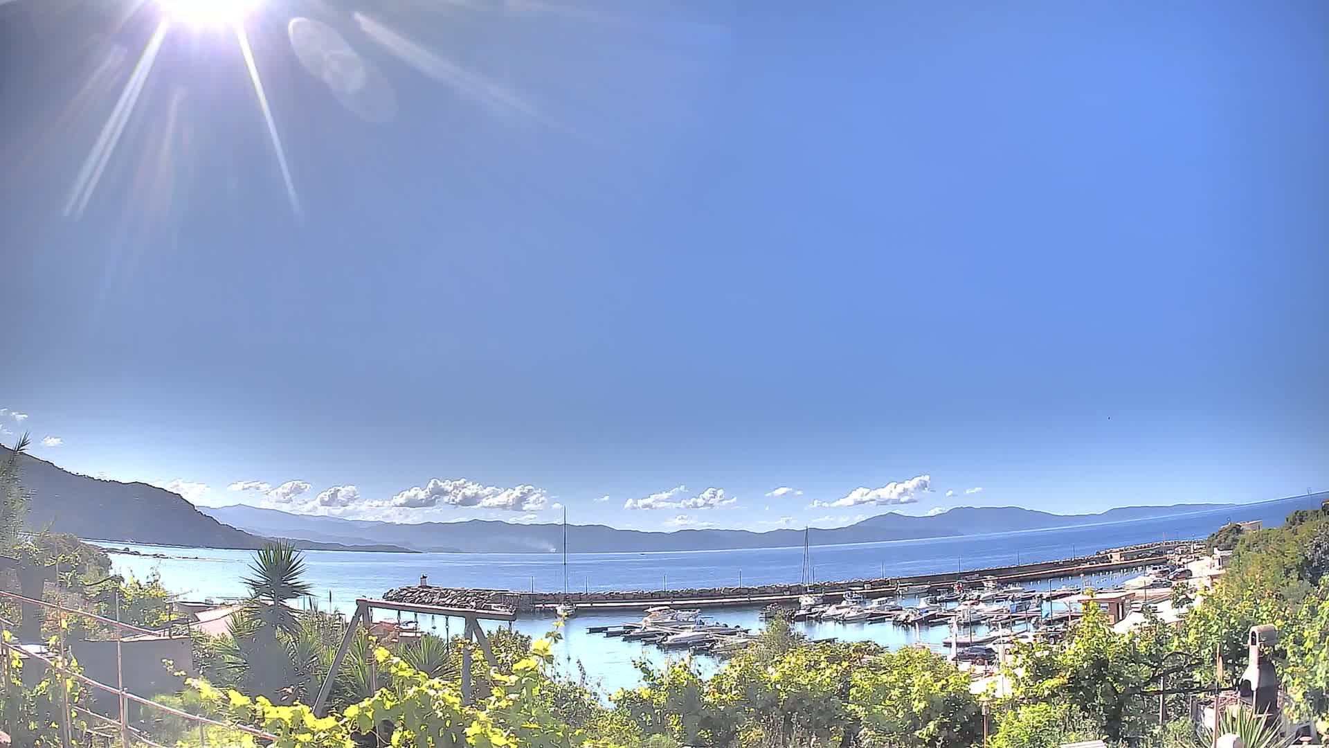 A sunny day reveals a harbor filled with boats, nestled between hills and a calm blue sea under a mostly clear sky.