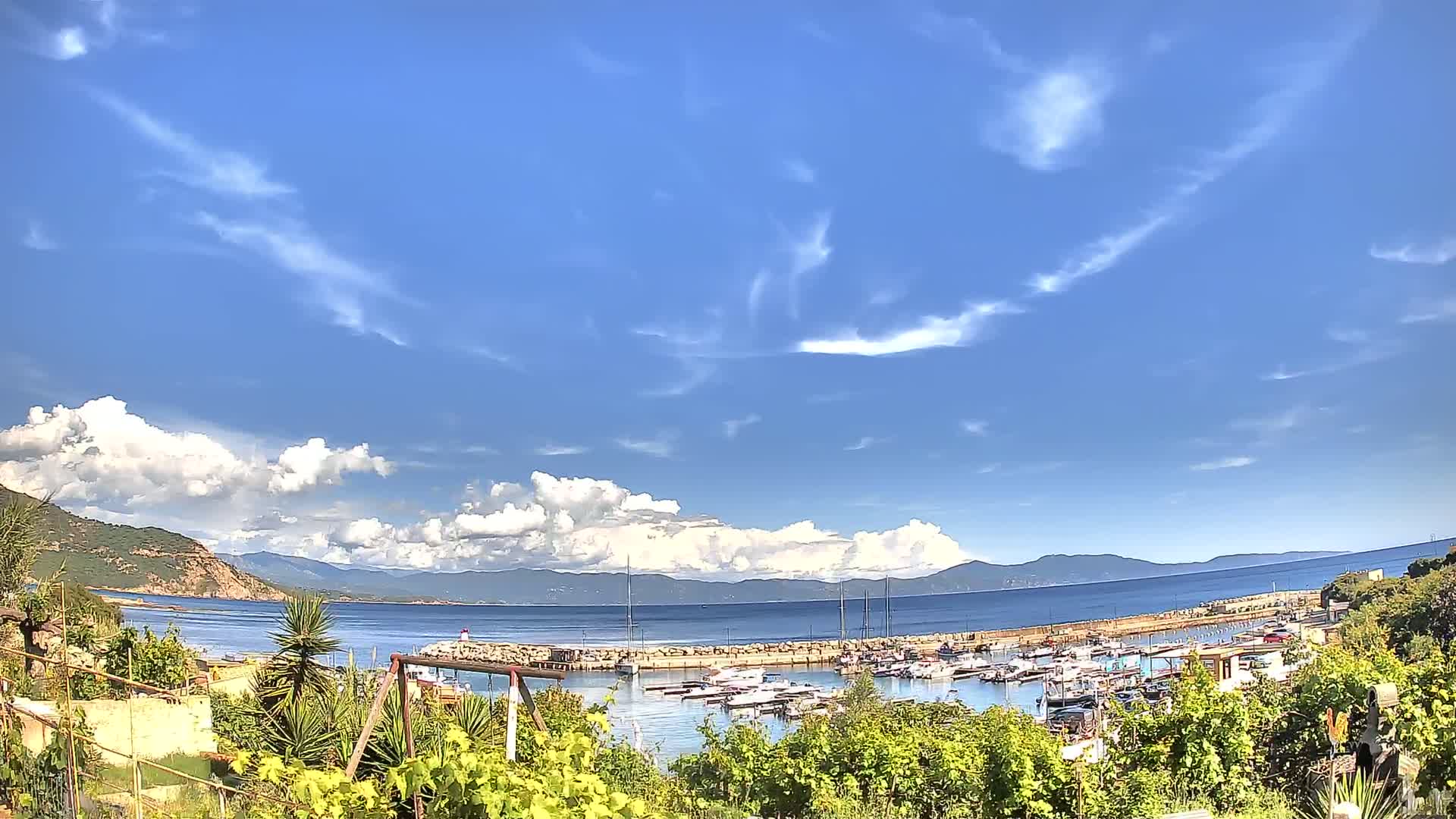 A partly sunny day shows a small marina filled with boats, nestled between a rocky shoreline and a calm sea with distant mountains under a partly cloudy sky.