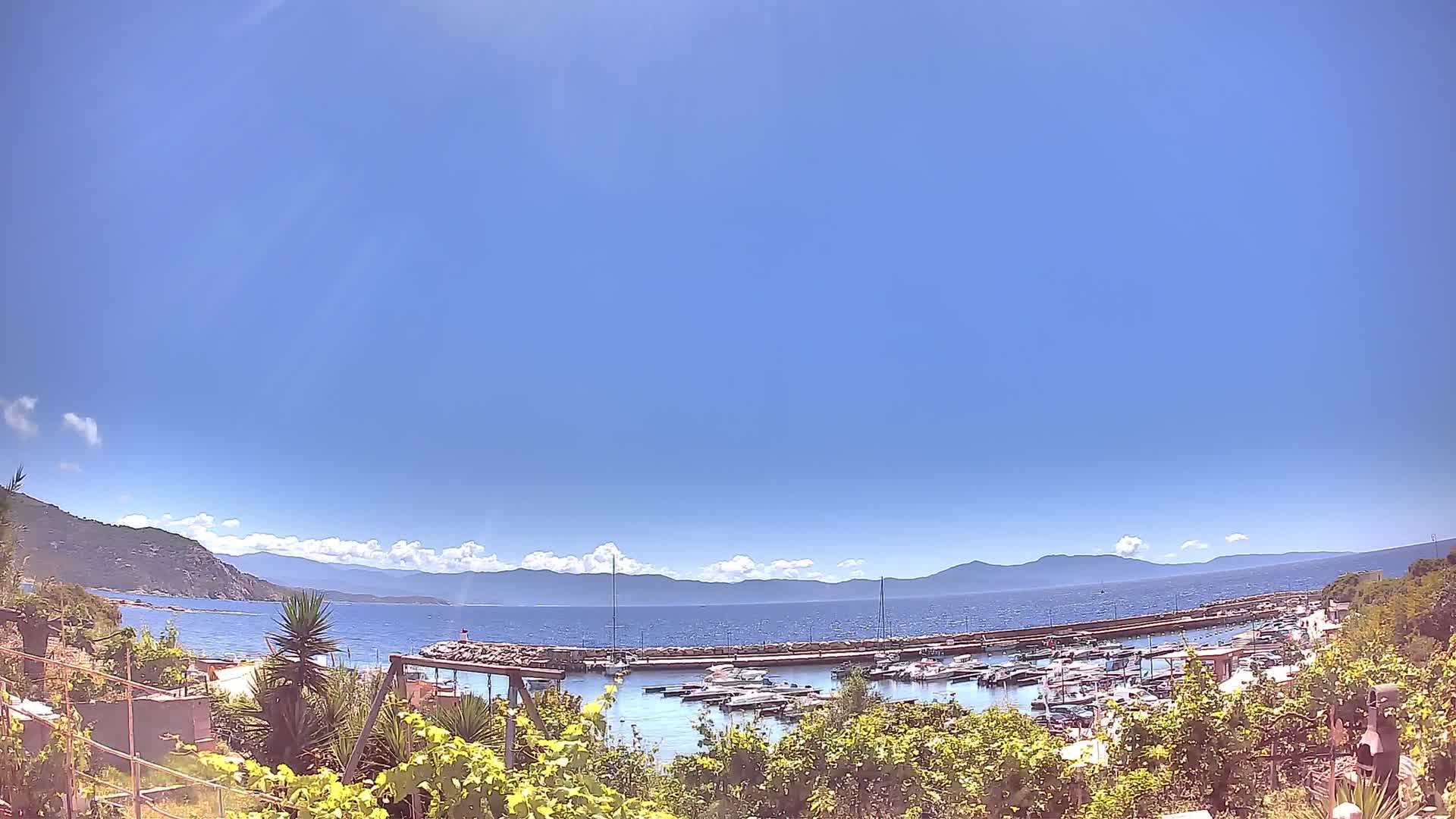 A sunny day reveals a harbor filled with boats, nestled between a verdant hillside and a calm sea under a mostly clear sky with a few scattered clouds.