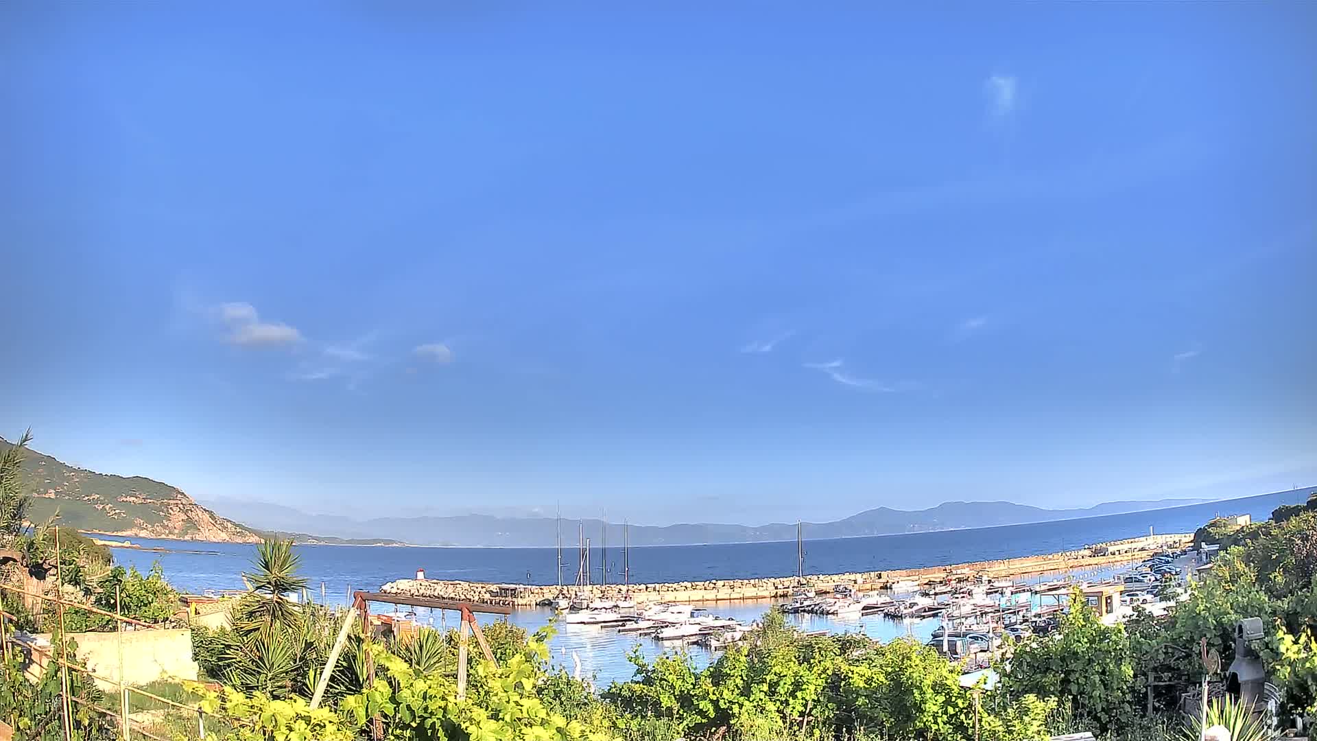 A calm, sunny day shows a harbor filled with boats nestled between a rocky coastline and distant mountains.