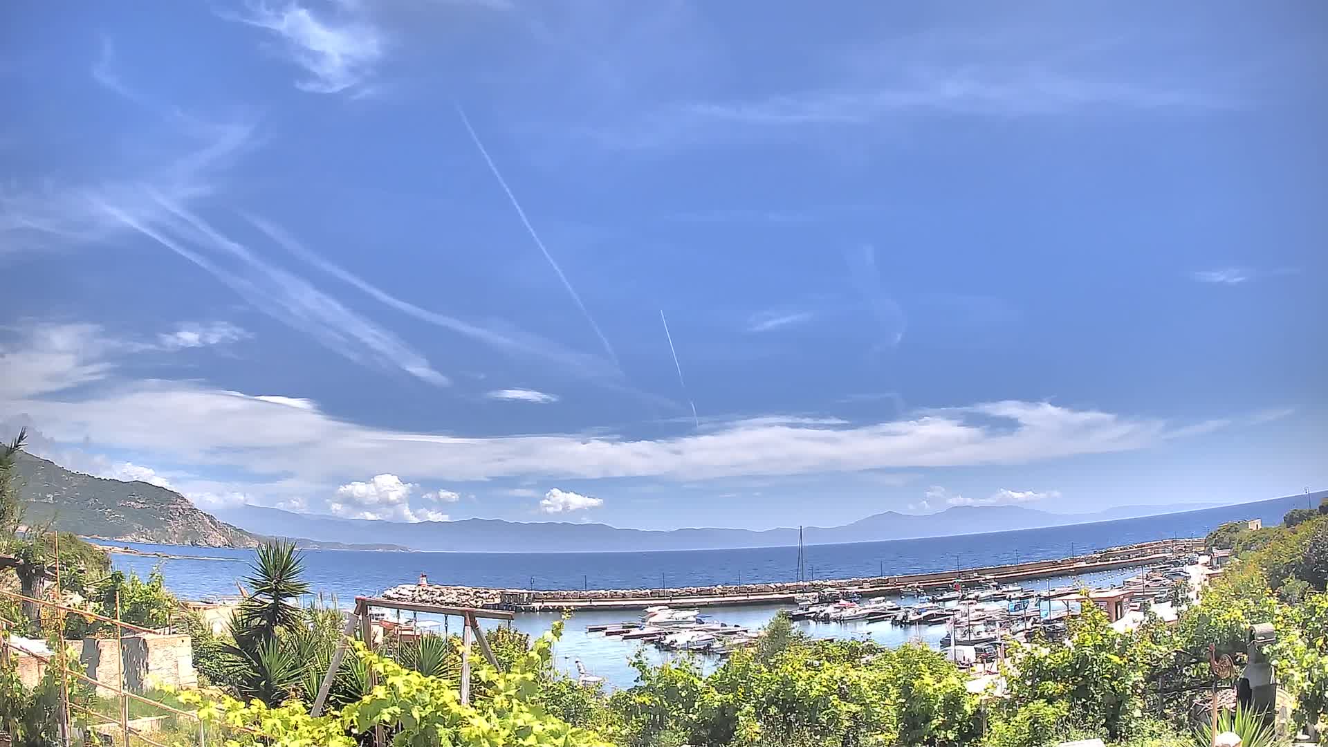 A marina filled with boats sits below a mostly sunny sky with scattered clouds and contrails, overlooking a calm sea and distant mountains.