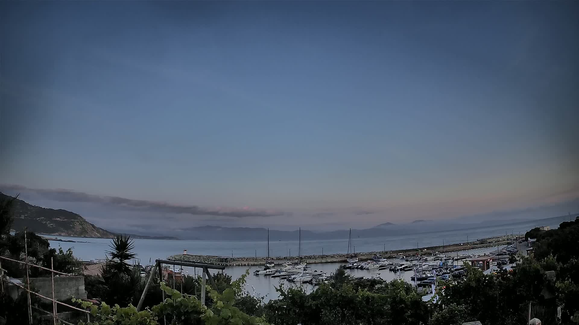 A calm, twilight scene shows a marina filled with boats, nestled between a verdant hillside and a hazy, mountainous coastline under a mostly clear sky.