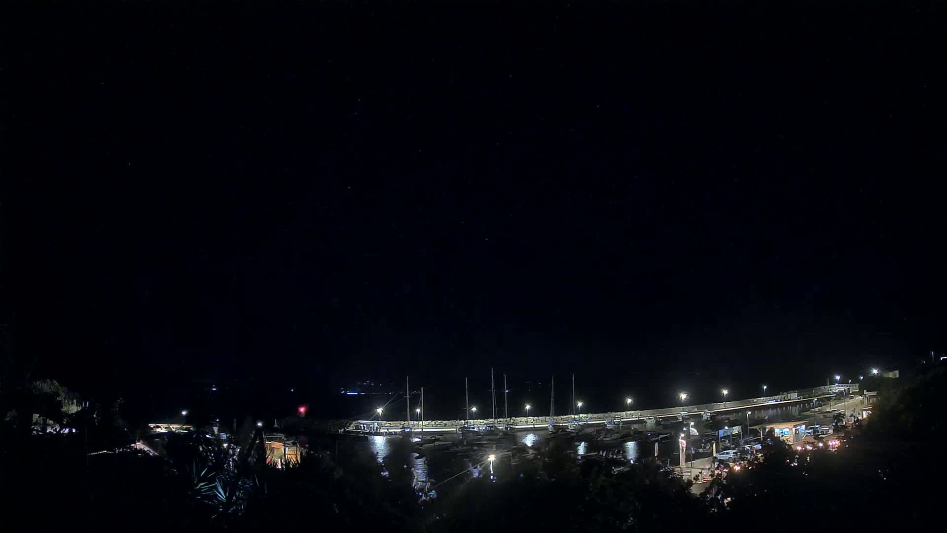 A nighttime view of a marina with boats docked, illuminated by lights, under a clear, dark sky.
