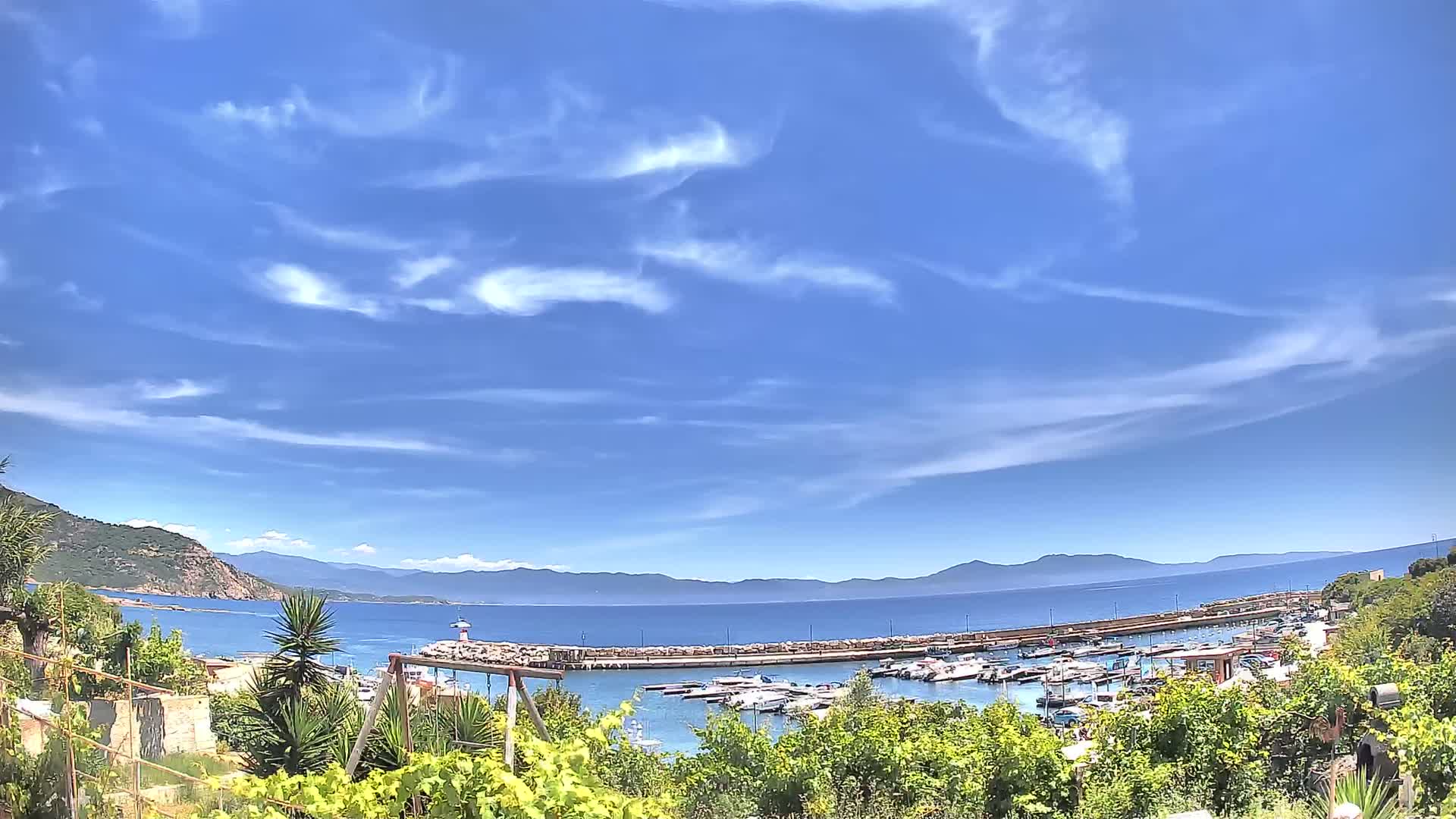 A small harbor filled with boats is nestled between green hills under a bright blue sky with wispy clouds.