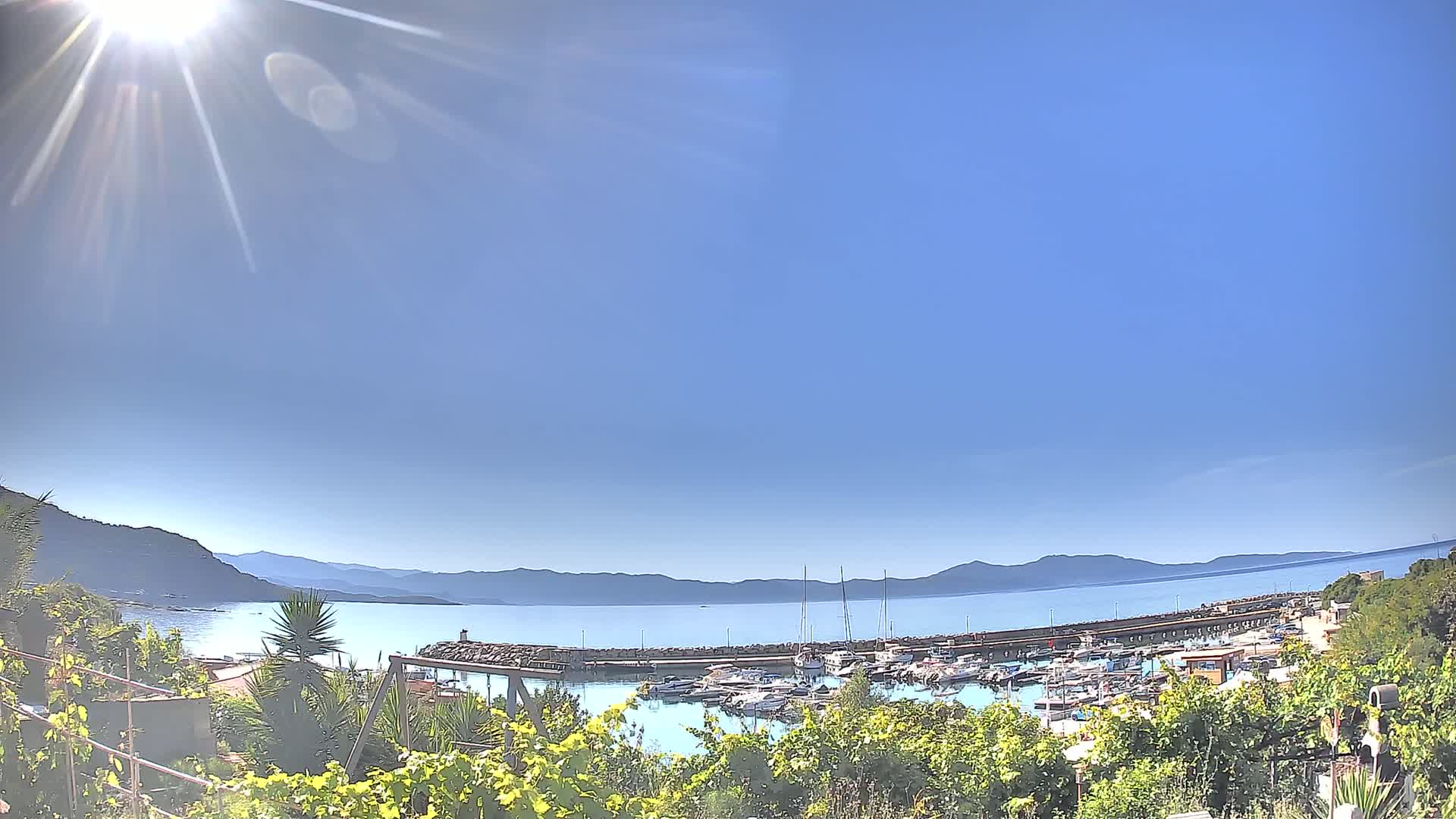 A sunny day overlooks a calm bay filled with boats, docked at a harbor wall, with mountains in the distance.