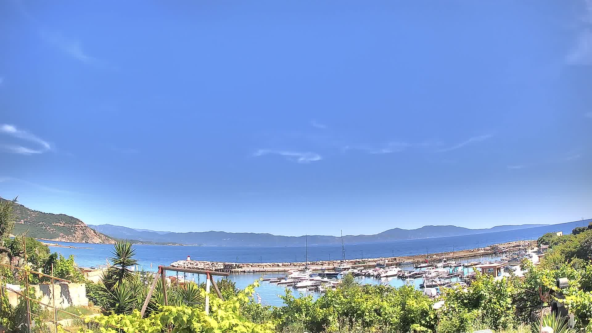 A small marina filled with boats is nestled between a rocky shoreline and distant mountains under a clear blue sky with a few wispy clouds.