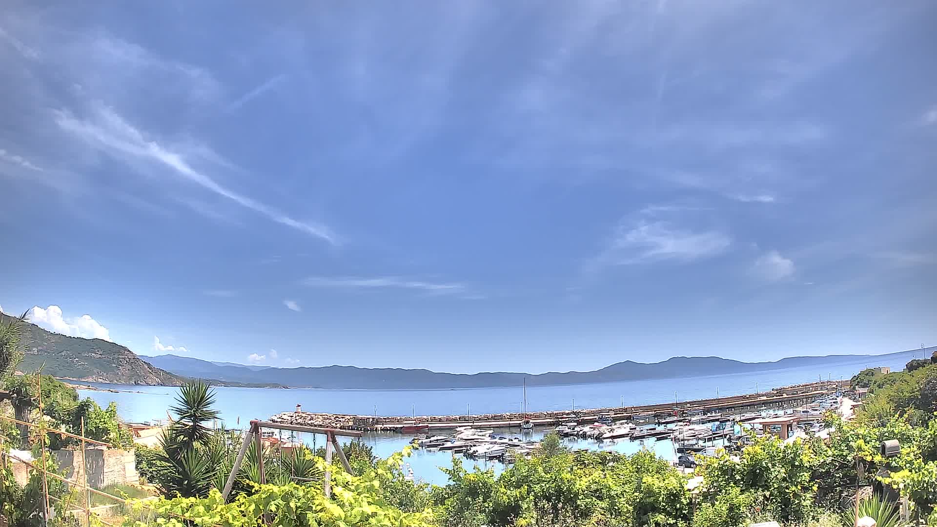 A marina filled with boats is nestled in a calm bay, under a mostly clear blue sky with a few wispy clouds, and backed by distant mountains.