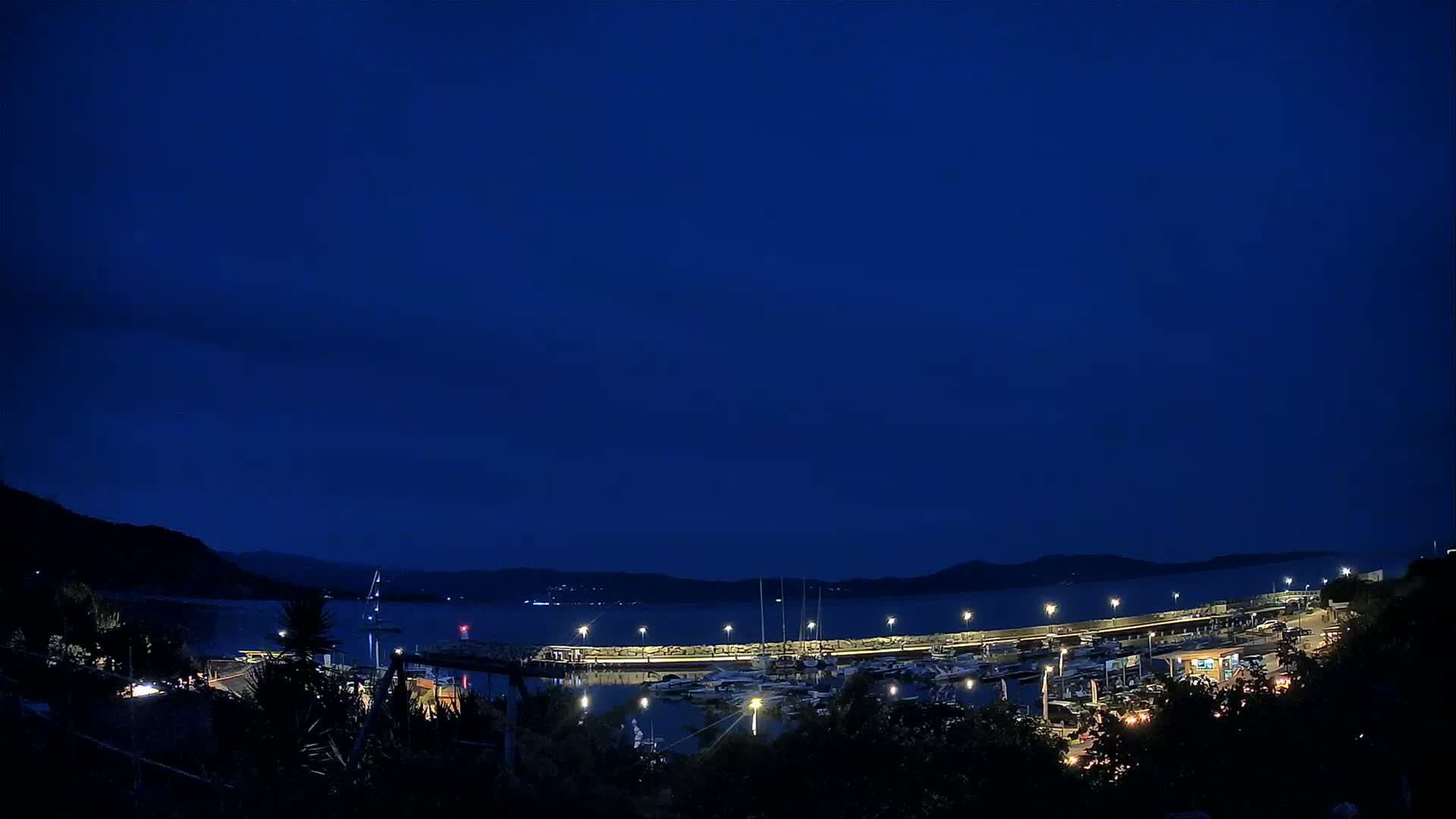 Under a dark blue night sky, a marina filled with boats is illuminated, nestled between hills overlooking a calm body of water.