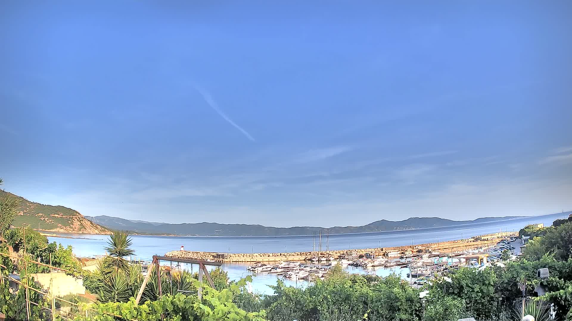 A clear blue sky overlooks a calm bay filled with boats, a small town, and hills in the distance.
