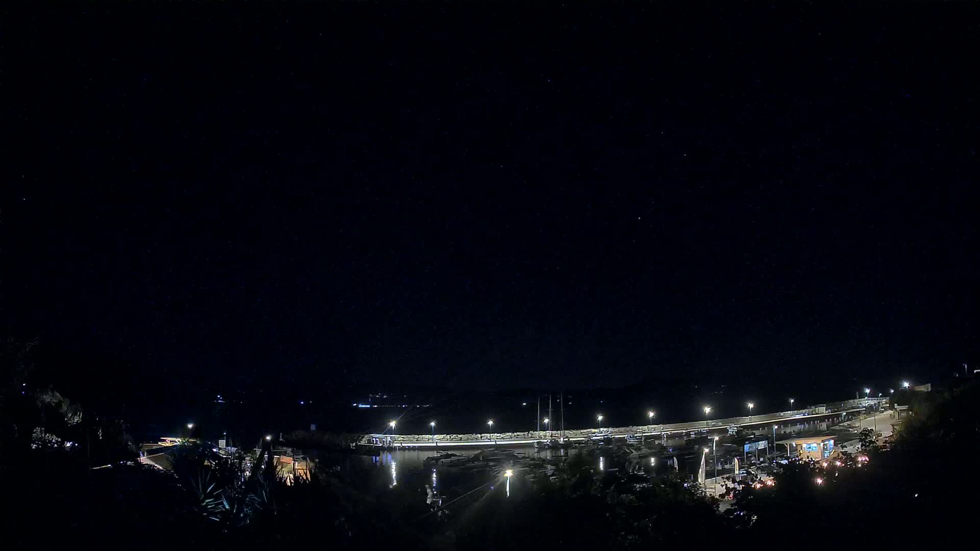 A nighttime view of a harbor filled with boats, illuminated by lights under a clear, starry sky.