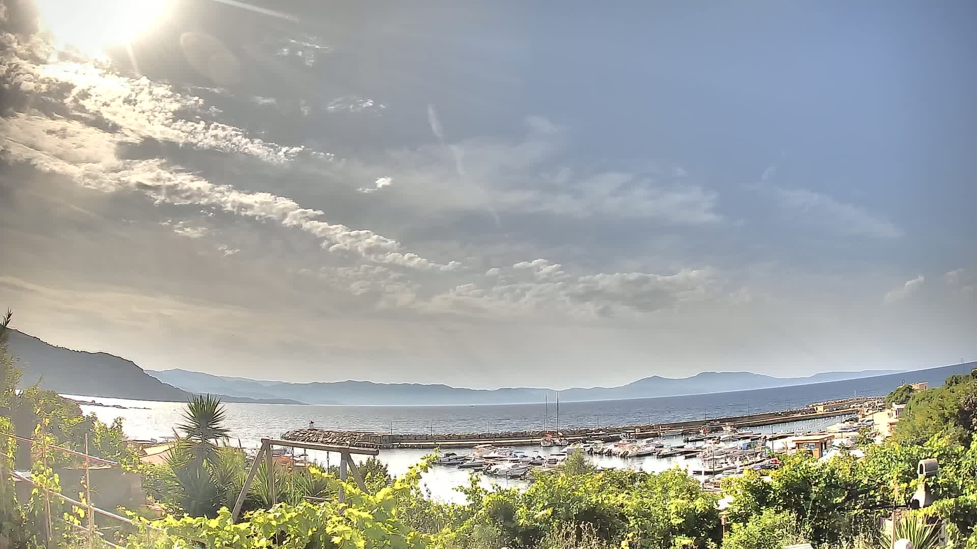 A sunny day with partly cloudy skies overlooks a harbor filled with boats, situated between a coastline and distant mountains.