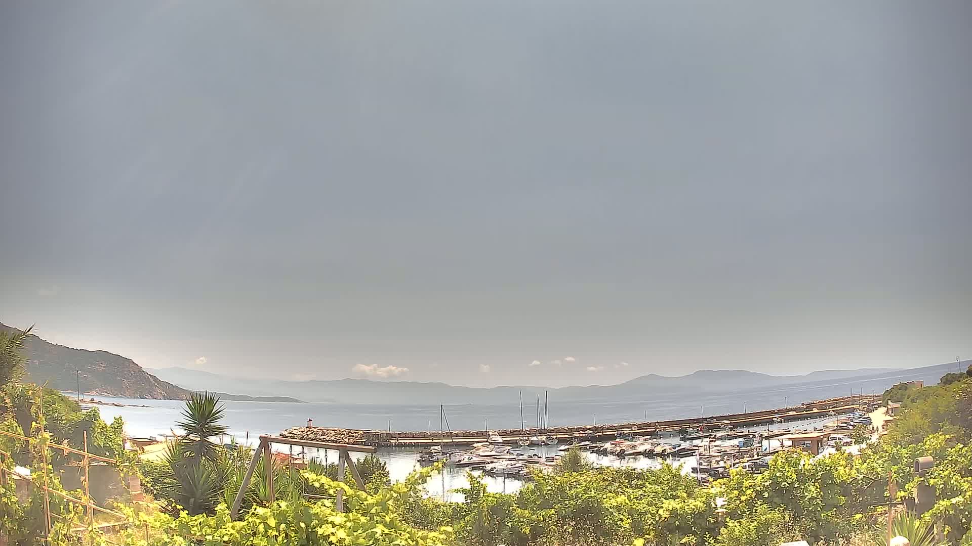 A marina filled with boats is seen from above under an overcast sky, nestled between a verdant hillside and a calm sea with distant mountains.
