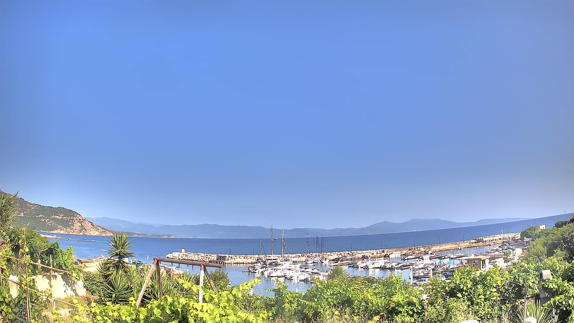A marina filled with boats is nestled between a verdant hillside and a calm, blue sea under a clear, bright sky.