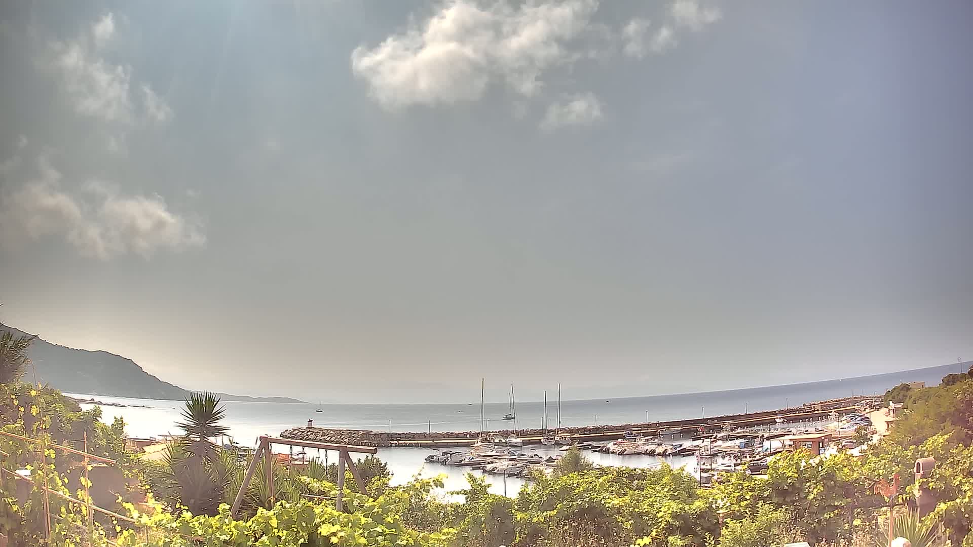 A partly cloudy day reveals a calm ocean bay with a marina filled with boats, viewed from a hilltop covered in lush green vegetation.
