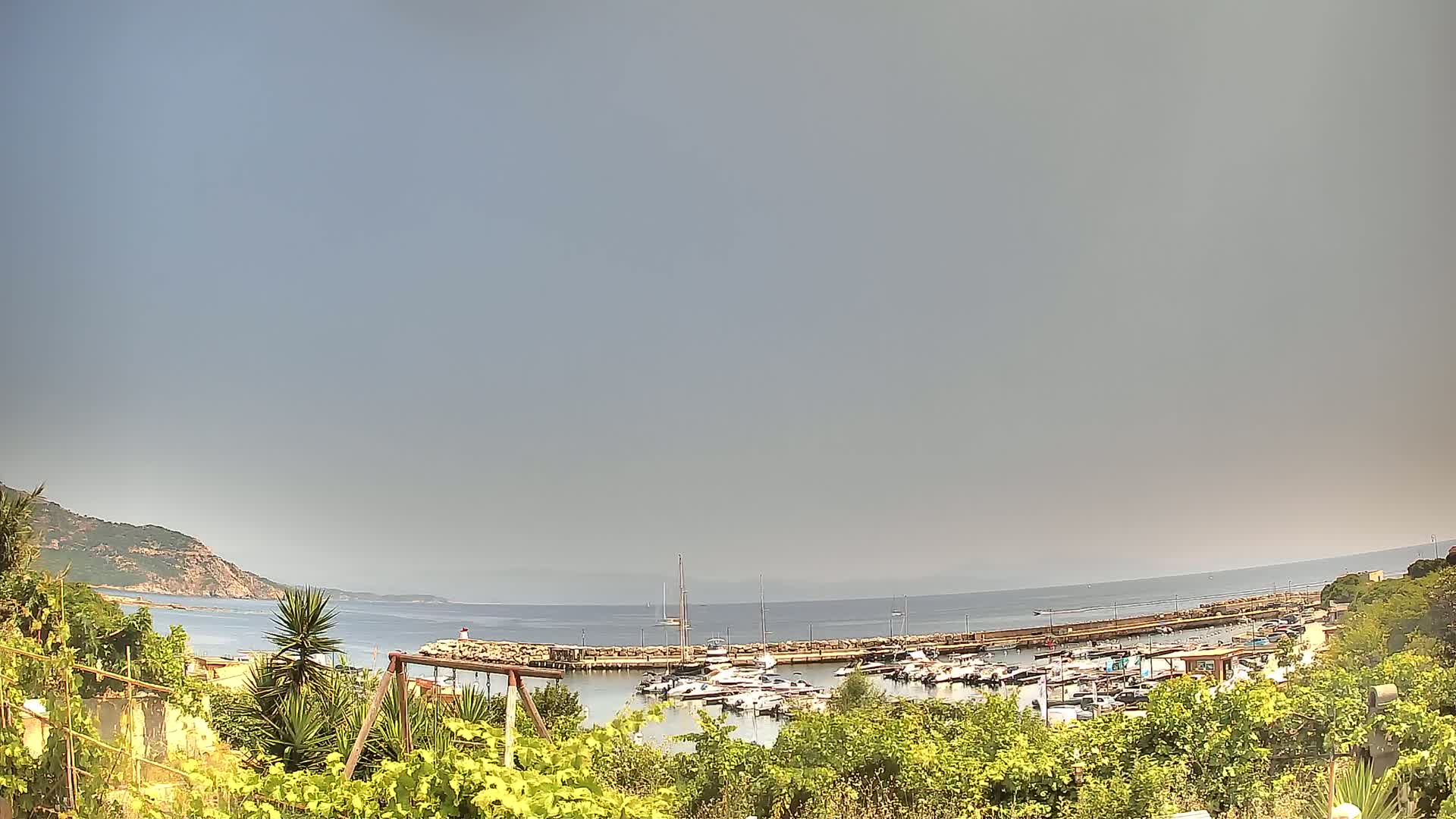 A hazy, sunny day overlooks a calm bay with a marina filled with boats, nestled between a green hillside and the ocean.