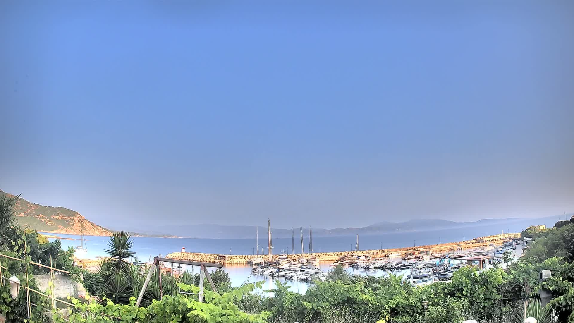 A calm, clear day reveals a marina filled with boats nestled between a rocky shoreline and a calm sea, all viewed from behind lush greenery.