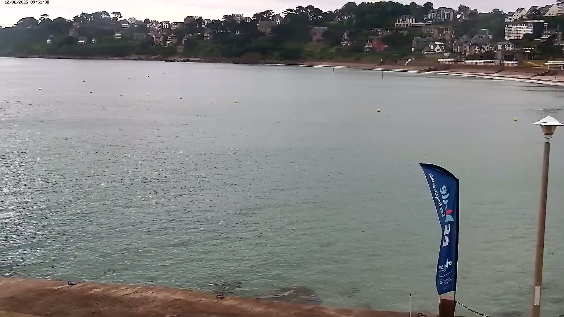 A calm, overcast day at a beach with a body of water, showing a shoreline with buildings in the background.