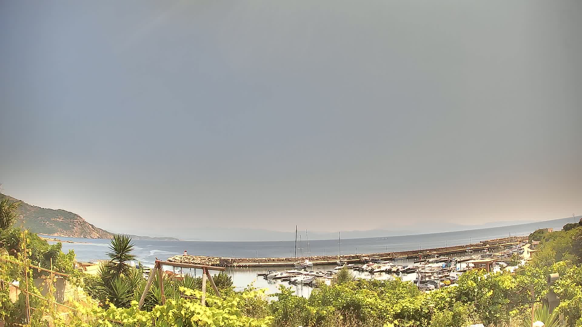 A hazy sky overlooks a calm sea harbor filled with boats, nestled between green hillsides and vegetation.