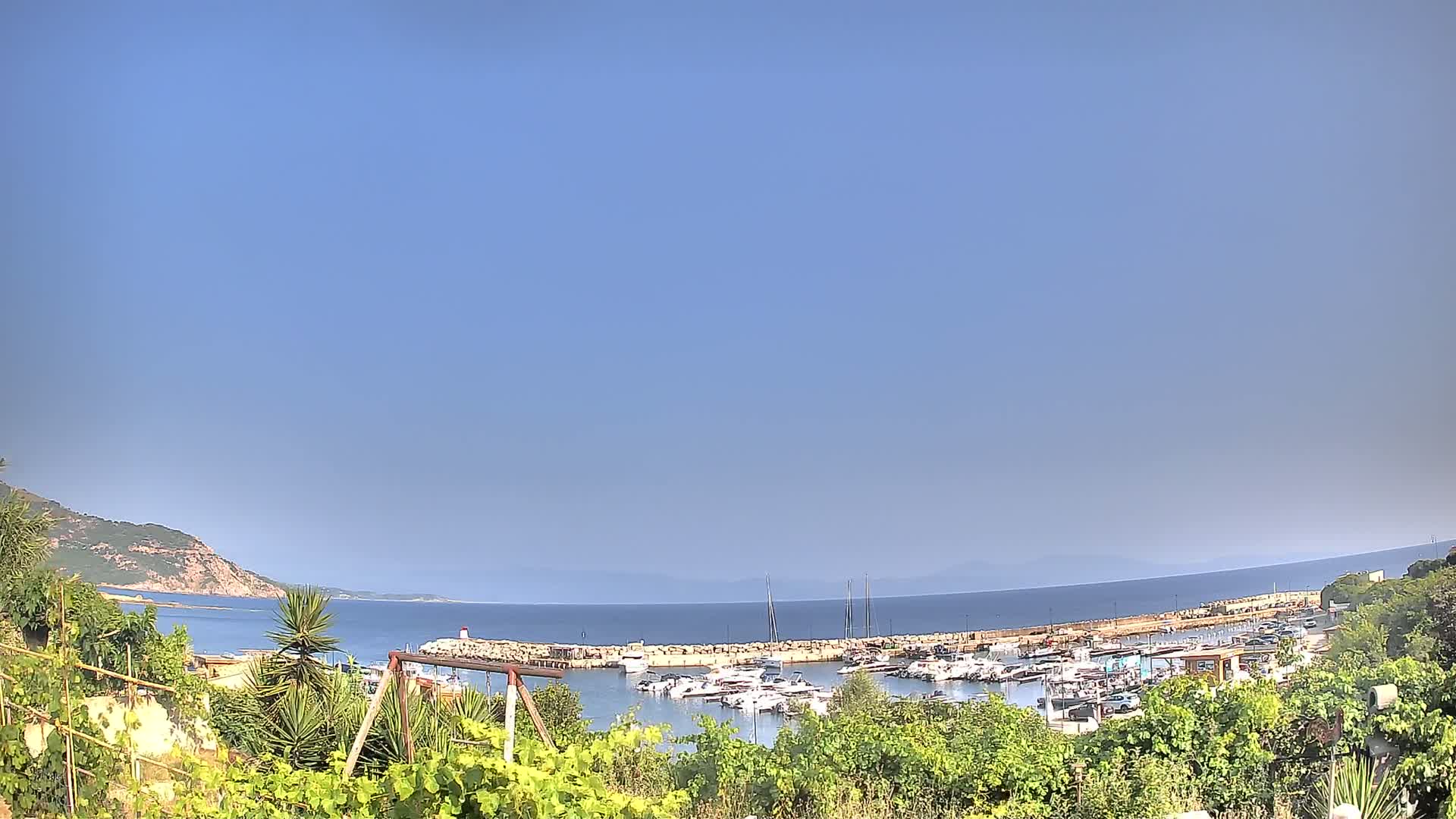 A calm, clear day reveals a marina filled with boats nestled between a rocky hillside and a calm blue sea.