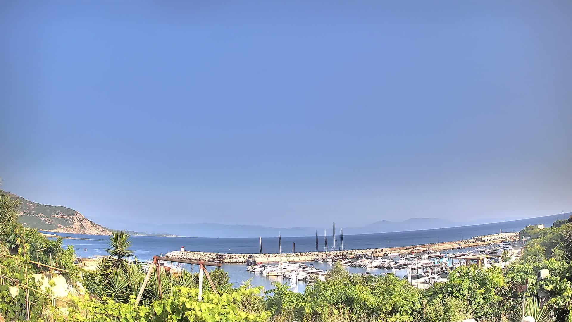 A marina filled with boats is nestled between a rocky hillside and calm, clear ocean under a bright blue sky.