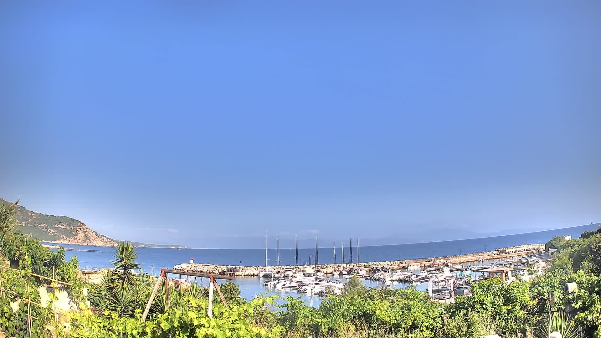 A marina filled with boats is nestled between a calm, blue sea and a verdant hillside under a clear, bright blue sky.