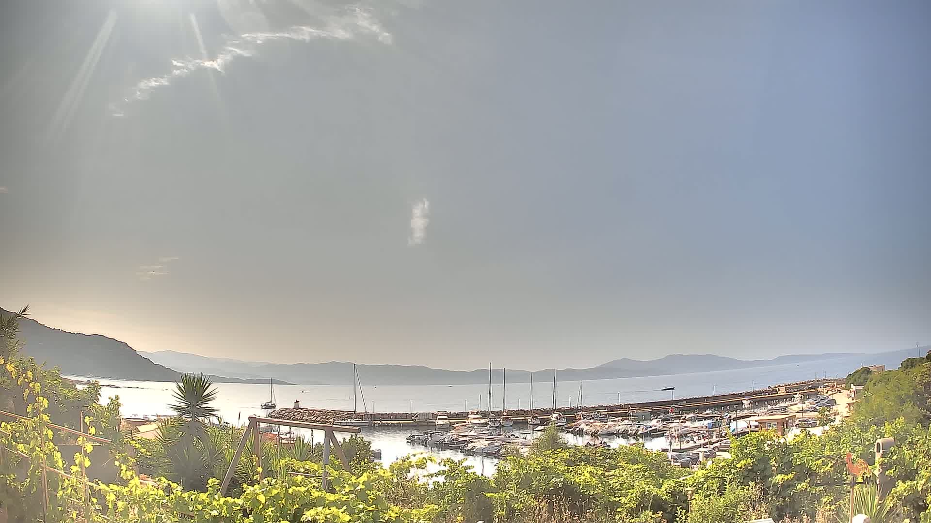 A sunny day reveals a marina filled with boats, nestled between a hillside and calm, blue water, extending to distant mountains under a mostly clear sky.