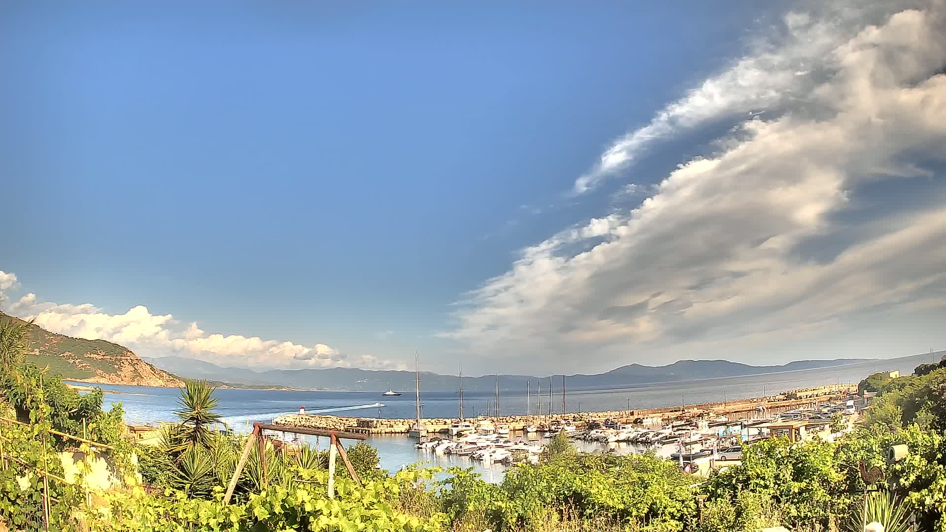 A partly cloudy sky overlooks a calm bay filled with sailboats and yachts, nestled between hillsides and lush vegetation.