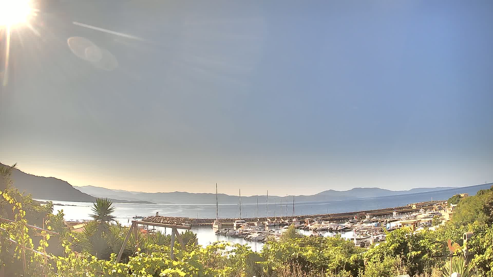 A sunny day reveals a harbor filled with boats, nestled between hills and a calm body of water under a clear sky.