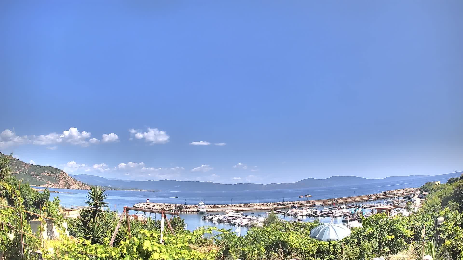 A calm, sunny day reveals a marina filled with boats nestled between a verdant hillside and a clear blue sea, backed by distant mountains under a mostly clear sky.