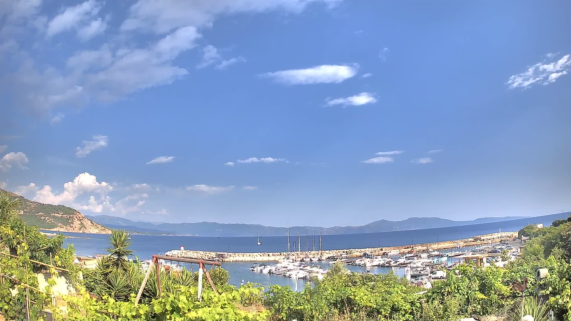A marina filled with boats is nestled between a hillside and calm blue water under a partly cloudy sky.