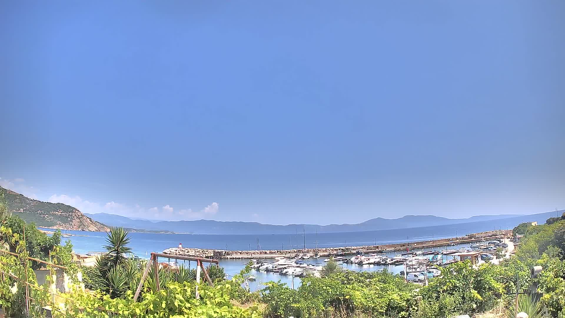 A marina filled with boats is nestled between a rocky shoreline and calm, blue water under a clear sky, with mountains visible in the distance.