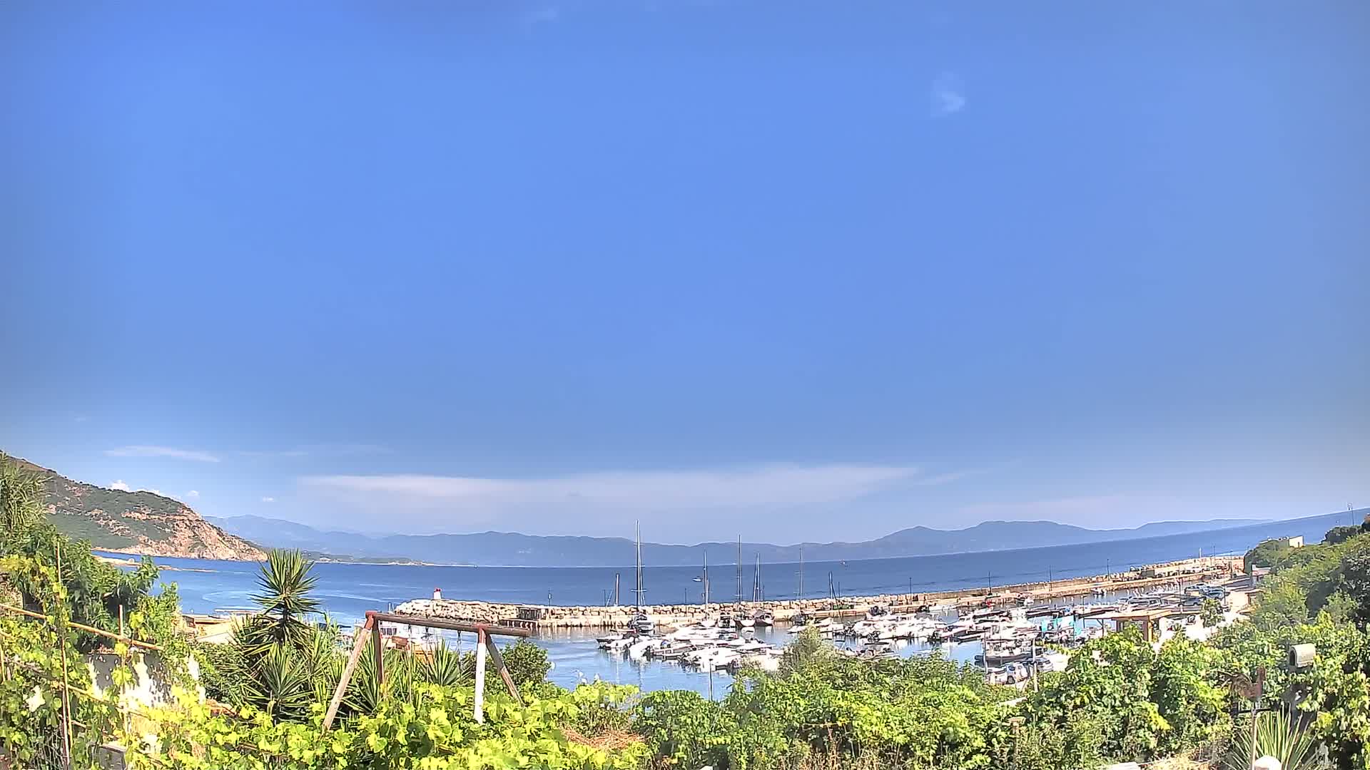 A marina filled with boats is nestled between green hills and a calm, blue sea under a clear sky.